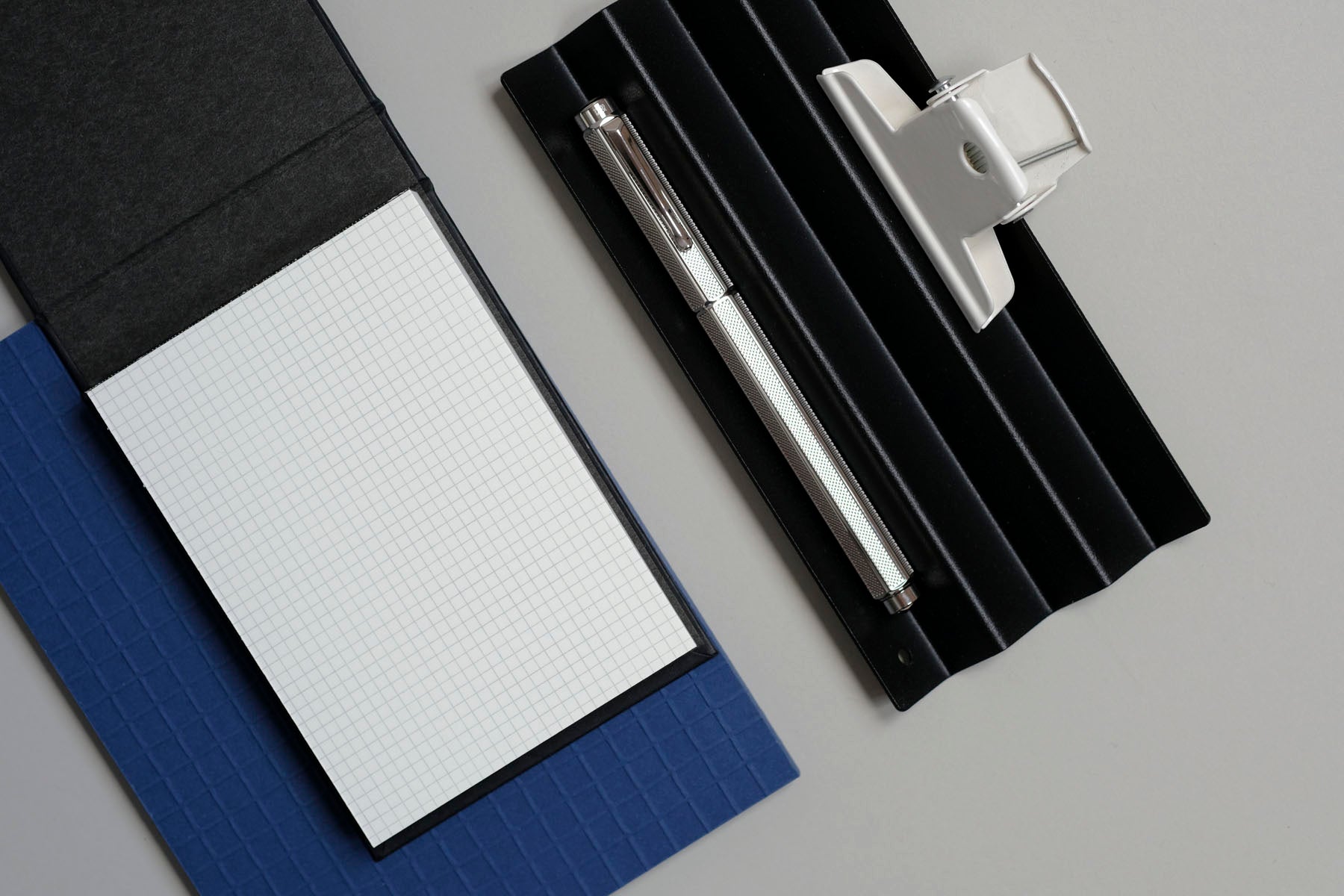 Podstawka na przybory NAKA w kolorze czarnym, a na niej pióro wieczne Ecridor Milanese Mesh i biały spinacz. Obok Work pad w kratkę i notatnik Martina w kolorze niebieskim.